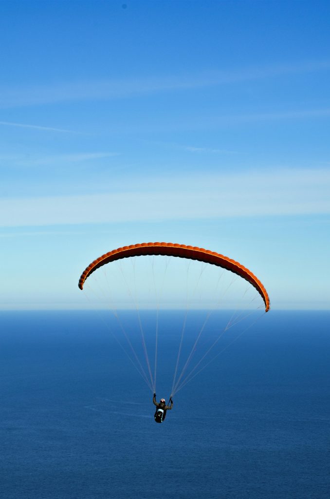 A person paragliding with an orange chute over a vast ocean under clear blue skies.