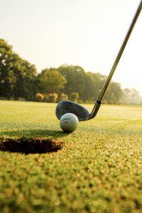 Closeup of golf ball placed on green grassy court against golf club under clear blue sky on sunny day